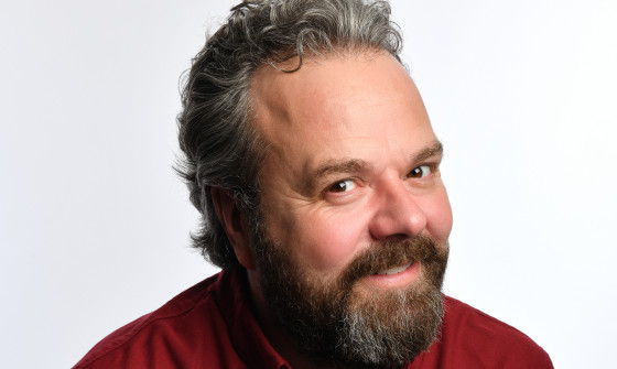 Hal Cruttenden smiles at the camera as he sits in a burgundy shirt.