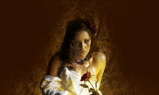 A young woman sits in a pile of straw in a barn with a white prairie dress on holding a red rose.