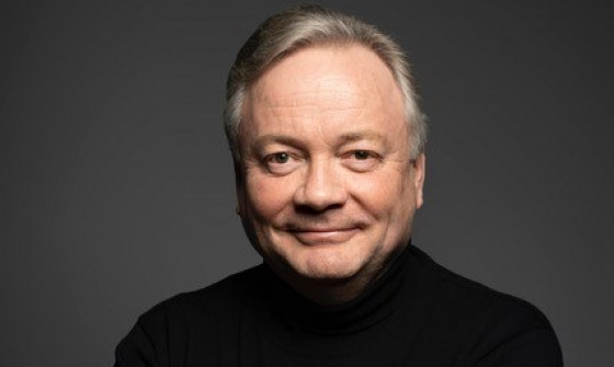 David Hill stands facing the camera with his arms crossed, in a black jumper with a dark background