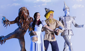 The Lion, Dorothy, Scarecrow, and Tinman are all linking arms as they skip across a green hill