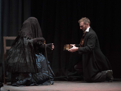 A man kneels before a veiled, cloaked figure and offers her a wooden box