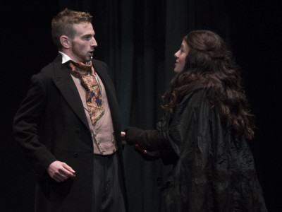 A cloaked woman approaches a man in a waistcoat, who looks taken aback by her presence.
