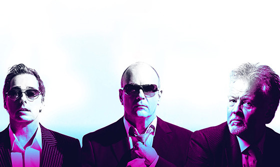 The three members of Go West sit in front of a white background with a blue and pink filter