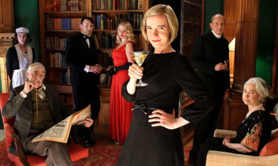 Lucy Worsley stands in a black dress wearing a martini glass. She is in a room of people in classic "murder mystery" attire.