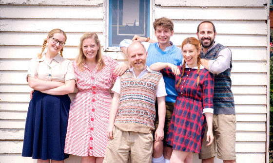 The cast of Bumper Blyton stand in front of a tiled house in costume (sweater vests, button down dresses and high socks).