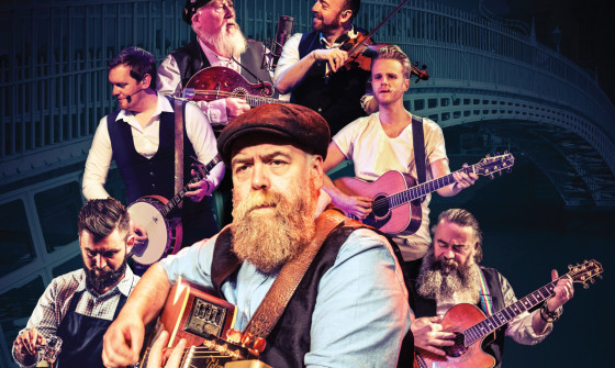 7 musicians perform with their instruments. A bearded guitarist stands at the front as the main focus of the image.
