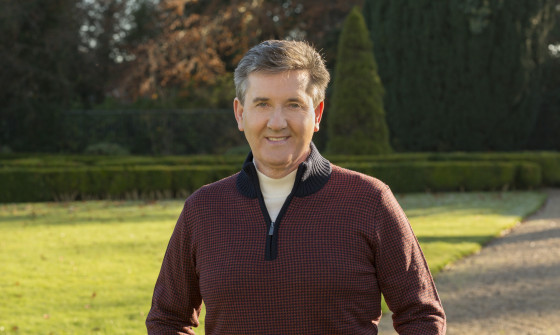 A mid waist shot of Daniel standing in a park. He is wearing a burgundy zip up jumper