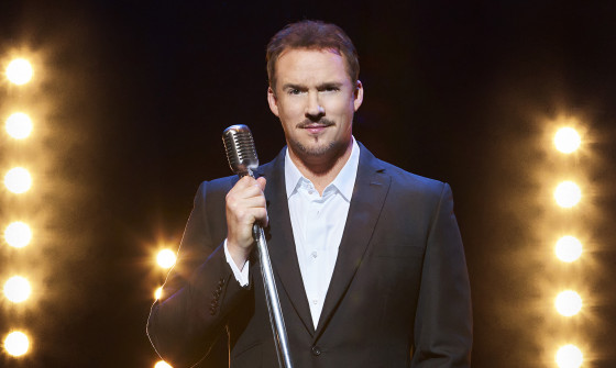 Russell Watson stands on stage holding a microphone