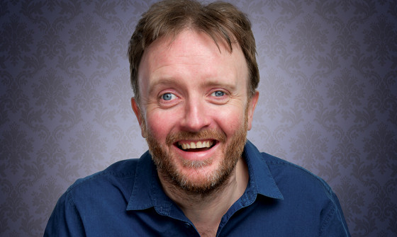 Chris McCausland smiles at the camera as he sits in a blue shirt