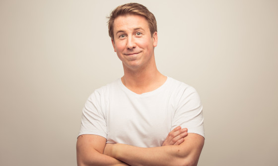 Carl Hutchinson stood smiling to the camera with his arms crossed, in a white t-shirt, with a neutral background