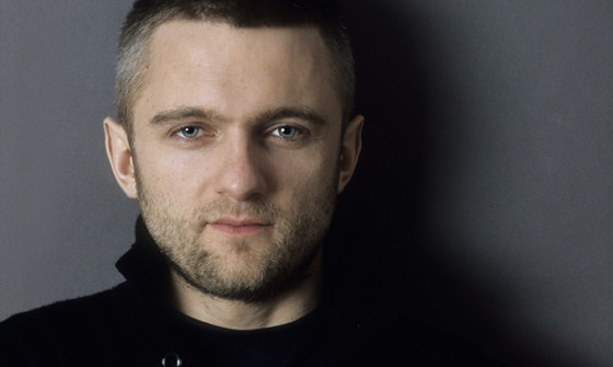 Kirill Karabits stands against a grey wall. His arms are crossed in front of him. He is wearing a black jumper