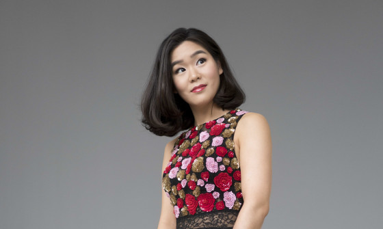 A full body shot of Esther Yoo holding her violin. She is wearing a dress. The top half has a floral pattern, the long skirt is black