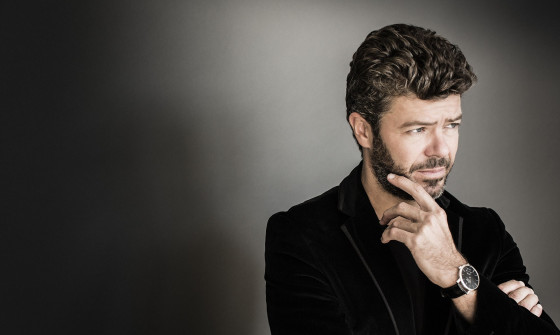 An upper shot of Pablo Heras-Casado. He is wearing a black blazer. His arms are crossed and his hand is on his chin
