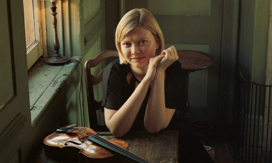 Alina Ibragimova is sat behind a small wooden table, her violin is placed on top. She is resting her elbows on the table