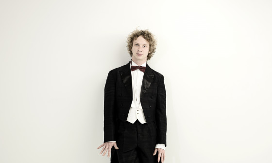 Santtu-Matias Rouvali is wearing a black tuxedo. He is standing against a white wall