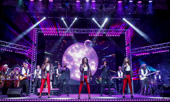 The cast of Lost in Music stand on stage in matching red trousers and black jackets in front of a large glitter disco ball.