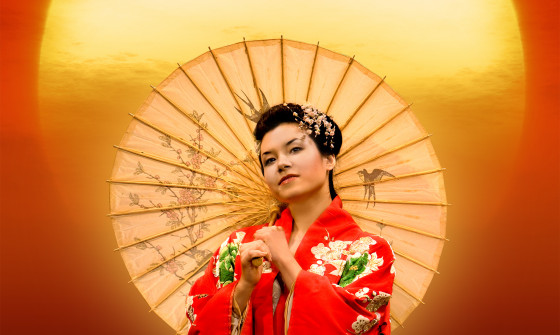 A woman stands in a striking red traditional kimono in front of a sunset. She holds a parasol which covers half of the sun.