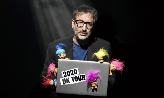 David Baddiel sits at a laptop in darkness with a spotlight on him. Six little colourful Troll dolls climb up and on his laptop.