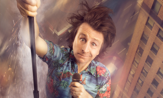 Milton Jones hangs out of a helicopter above a city. He is holding a microphone.