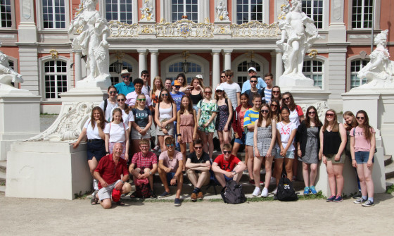 A group of students stand in front of some statues and a regal home.