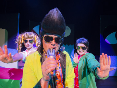 A man with a quiff sings into a microphone with two backing dancers