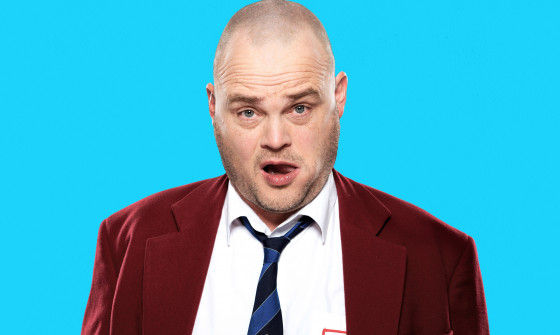 Al Murray stands in a blazer and tie in front of a blue background