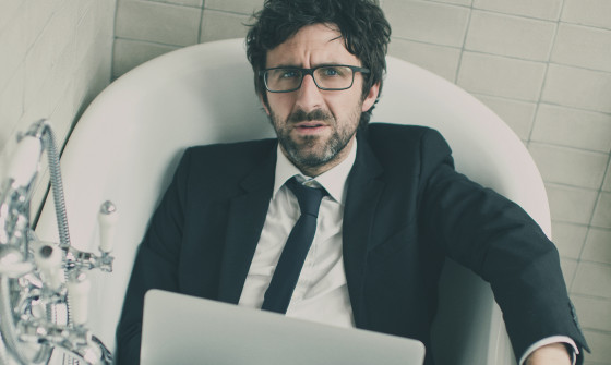Mark Watson sits in a bathtub in a suit with a laptop on his lap.