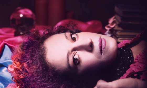 Kate Rusby lies on a blue satin sheet looking at the camera. She has pink hair and a pink dress.