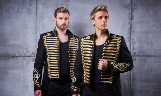 Jonathan Ansell and Jai McDowall stand in military drummer jackets against an industrial wall