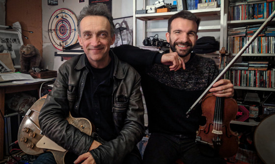 Justin and Mauro sit in front of a bookshelf, holding their intruments